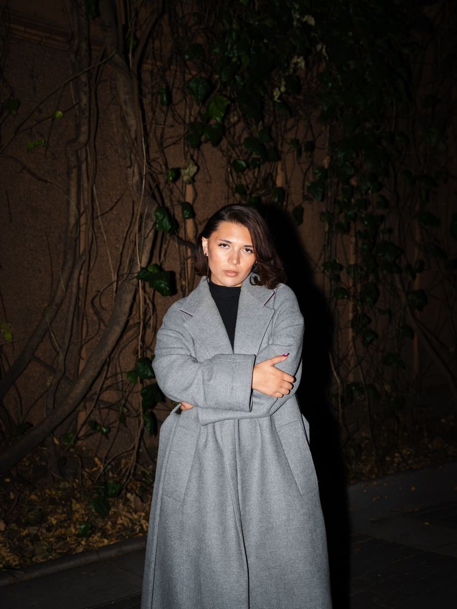 Night flash portrait in front of an ivy wall, long grey coat, arms crossed.