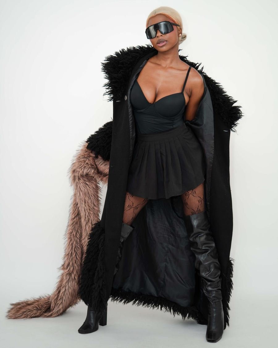 Studio full length, black coat with fur trim, visor sunglasses and thigh boots.